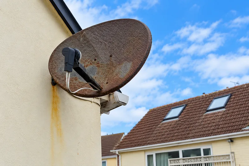 Heavily rusted satellite dish and mounting bracket on an exterior wall showing corrosion and weather damage