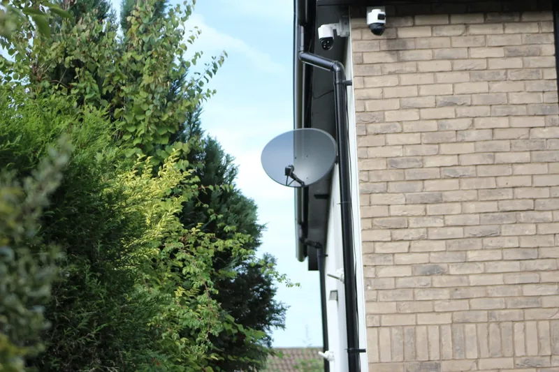 Satellite dish mounted on a brick house wall above surrounding trees